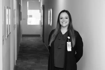 Traci is standing in a hallway, smiling, with her hands resting behind her back. She's wearing a dark cardigan and her hospital ID badge is clipped to her shirt.
