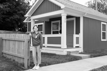 Emily rests her right elbow on a short wooden fence. She stands in front of a quaint one-story residence with bright white trim.